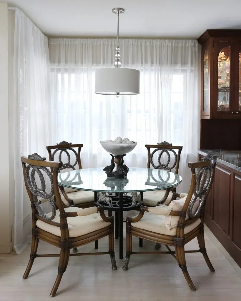 Glass dining table with carved wood chairs, a decorative urn, and sheer curtains